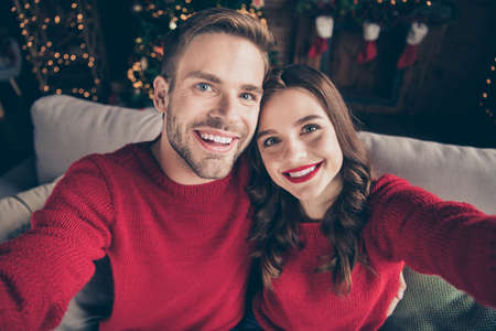 Photo of charming couple spending christmas together making selfies in decorated garland lights room near newyear tree indoors wear red pulloversの写真素材