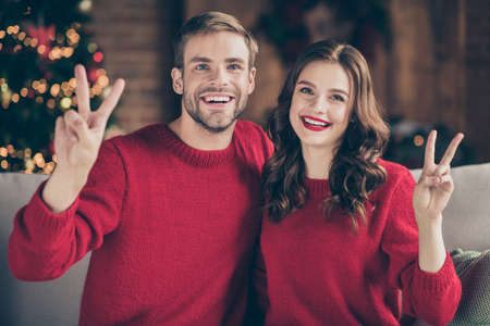 Photo of adorable couple spending x-mas eve in decorated garland lights room showing v-sign symbols sitting close on comfy sofa indoors wearing red sweatersの写真素材