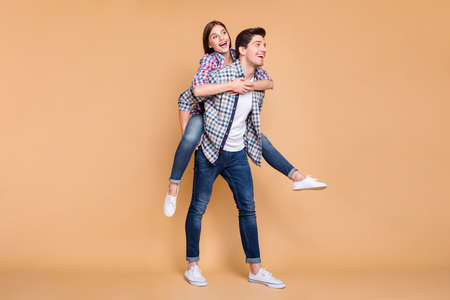 Full length body size turned photo of cheerful positive cute nice white couple wearing jeans denim with her having ridden his back and him carrying her isolated over beige pastel color backgroundの写真素材