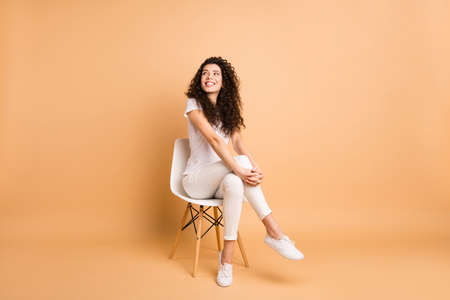 Full length photo of amazing lady looking interested empty space sitting comfy chair good mood inspired wear casual clothes isolated beige pastel color backgroundの写真素材