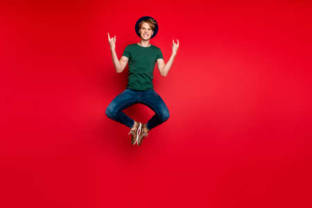 Full length photo of funny funky guy jump show horned sign enjoy rock concert feel candid content crazy wear headwear green t-shirt denim jeans sneakers isolated over red color backgroundの写真素材