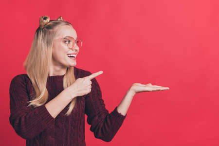 Photo of amazing lady holding open palm new sale price product indicating finger at it advising good quality wear specs knitted jumper isolated on pastel red color backgroundの写真素材