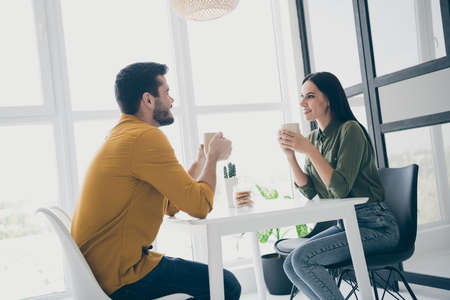 Profile photo of handsome guy and his pretty lady looking tender eyes drinking hot beverage sitting chairs opposite in stylish interior light cafeteria indoors wear casual clothesの写真素材