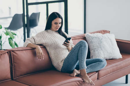 Profile side photo of concentrated girl use her smartphone have inline conversation with friends write sms sit on brown leather divan in house indoors wear knitted sweater denim jeansの写真素材