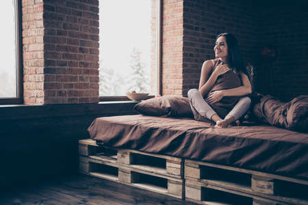 Nice attractive charming cute pretty lovely winsome cheerful cheery girl sitting in bed hugging pillow enjoying perfect weekend at industrial brick loft modern style roomの写真素材