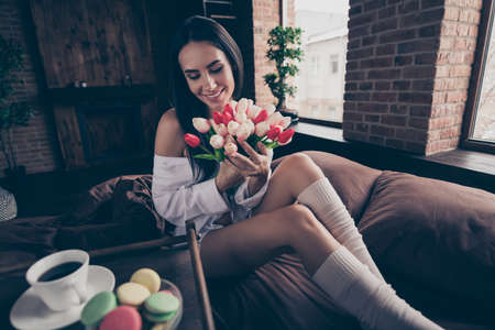 Portrait of her she nice attractive charming pretty cheerful dreamy girl sitting on bed holding in hands enjoying smelling fresh tulips at industrial brick wood loft modern interior style houseの写真素材
