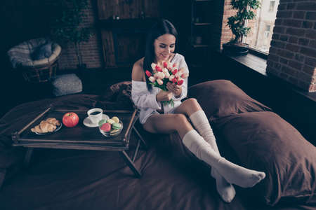 Portrait of her she nice attractive lovely charming cheerful cheery dreamy girl sitting on bed smelling fresh aromatic tulips at industrial brick wood loft modern interior style houseの写真素材