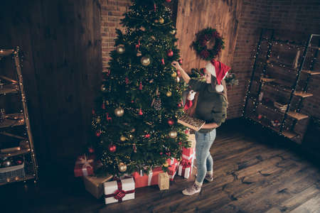 Top above high angle photo of positive brunette hair girl hand bauble on evergreen fir tree prepare for christmas celebration party wear santa claus hat in house with newyear decoration lights indoorsの写真素材