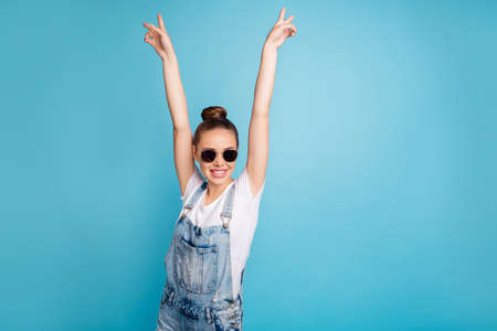Portrait of funny funky girl with eyeglasses eyewear raising v-signs wearing white t-shirt denim jeans overalls isolated over blue backgroundの写真素材