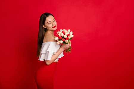 Portrait of cute youth enjoying, flowers aroma closing eyes wear white off-shoulders isolated over red backgroundの写真素材