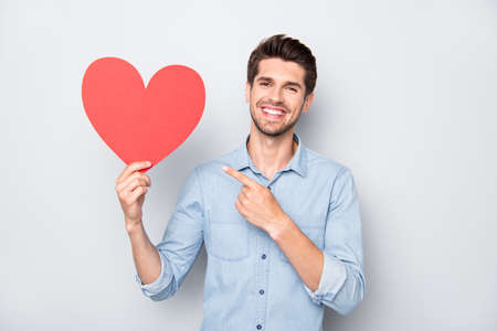 Portrait of charming man with positive emotions expression hold hand red big paper card heart present his gift for 14-february to girlfriend wear casual style clothes isolated grey color backgroundの写真素材