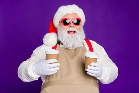 Close-up portrait of nice glad cheerful cheery bearded Santa Claus holding in hands two cups hot cacao sharing with friend isolated over bright vivid shine vibrant violet lilac backgroundの写真素材