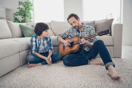 best teacher ever concept. Concentrated dreamy cheerful father showing his little son how to locate fingers on guitar neck sitting on floor barefoot indoors in flat roomの写真素材