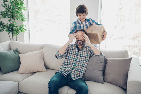 Portrait of two nice attractive cheerful cheery glad guys daddy pre-teen son hiding carton box for dads day occasion in light white modern interior house living-room indoorsの写真素材