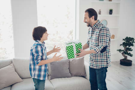 Profile side view portrait of two nice lovely cheerful cheery glad guys dad daddy giving festal box gift to pre-teen son in light white modern interior house living-room indoorsの写真素材