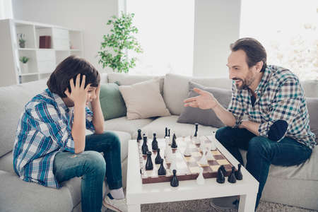 Portrait of two nice cheerful cheery glad guys dad and pre-teen son sitting on sofa playing chess...