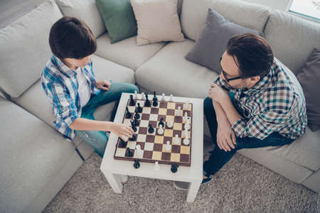 Top above high angle view of two nice attractive smart clever intellectual, guys dad and pre-teen son sitting on sofa playing chess moving pieces on board in light white modern interior house roomの写真素材