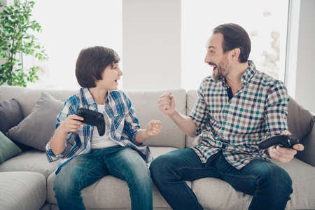 Portrait of two nice attractive satisfied cheerful cheery guys dad and pre-teen son sitting on sofa playing online game winner at light white modern style interior house living-room indoorsの写真素材