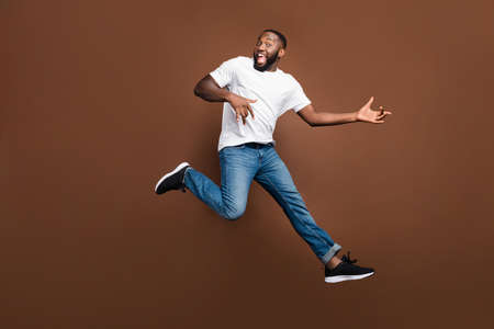 Full length body size photo of trendy stylish cheerful handsome guy running jumping playing guitar in air wearing white t-shirt casual jeans denim footwear isolated over pastel brown color backgroundの写真素材