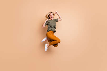 Full length profile photo of crazy lady jumping high competitive mood celebrating best victory triumphant wear green t-shirt yellow trousers isolated beige color backgroundの写真素材