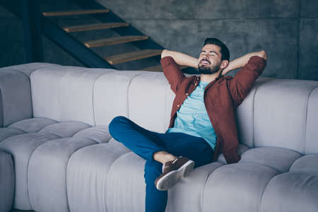 Photo of handsome guy in high spirits holding hands behind head sitting comfortable big couch sleeping relaxing saturday morning in modern flat loft indoorsの写真素材