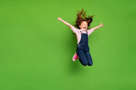 Full length photo of cheerful pretty little schoolchild jumping high rejoicing summer holidays hair flying wear casual denim overall pink shirt isolated green backgroundの写真素材