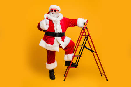 Full size photo of funky white beard hair santa claus in red hat climb ladder decorates christmas tree deliver wish dream gifts show thumb up approve x-mas celebration isolated yellow color backgroundの写真素材
