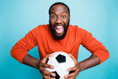 Portrait of cheerful excited mixed-race overjoyed ecstatic man holding, ball rejoicing in goal of his favorite team isolated over blue vivid color backgroundの写真素材
