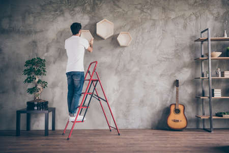 Full length rear behind view photo of mixed race arabian guy installing wall shelf standing on stepladder repair of new flat craftsman working hanging last shelf indoorsの写真素材