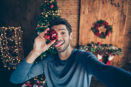 Photo of funny attractive fooling man covering his eye with christmas toy sticking tongue out taking...