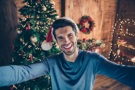 Self portrait of cheerful excited enjoying man having finished decorating his christmas tree and taking selfie on background of it smiling toothily in santa capの写真素材