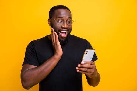 Photo of ecstatic overjoyed man browsing through his telephone rejoicing in receiving, positive notifications in t-shirt isolated vivid color yellow backgroundの写真素材