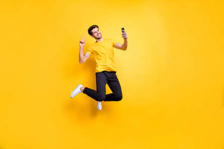 Full length photo of handsome guy jumping high holding telephone taking selfies, showing v-sign symbol wear casual t-shirt pants isolated yellow color backgroundの写真素材