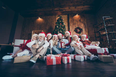 Photo of cheerful positive big family surrounded with gift boxed smiling toothily in front of christmas tree lights wreath in santa hat cap showing thumb upの写真素材