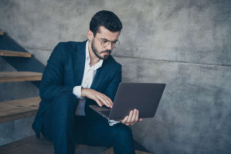 Portrait of his he nice attractive focused concentrated bearded guy financier holding in hands laptop browsing reading finance information sitting on stairs gray concrete wall backgroundの写真素材