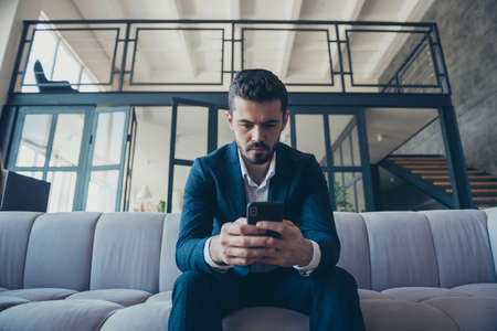 Low angle view photo of rich trendy handsome business guy sit sofa hold telephone read partners report project in apartment office industrial indoors wear formalwear costumeの写真素材