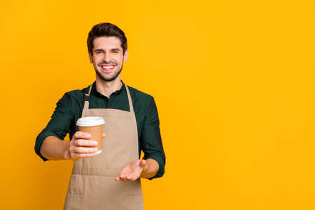 Here your latte. Portrait of positive cheerful guy student work part-time in coffee bar shop give to client paper card disposable mug wear stylish trendy outfit isolated over yellow color backgroundの写真素材