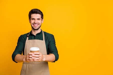 Photo of cheerful positive handsome attractive man holding paper cup with hands recommending you to taste this sort of coffee wearking apron isolated over yellow vivid color backgroundの写真素材