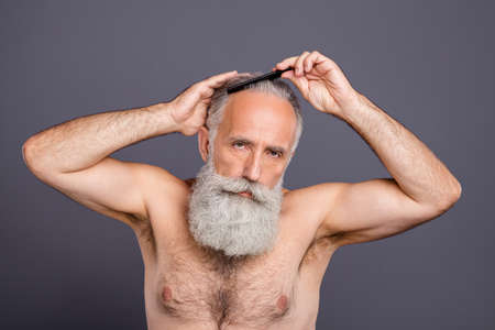 Photo of confident masculine handsome attractive mature old man combing his hair with hair brush isolated over grey color backgroundの写真素材