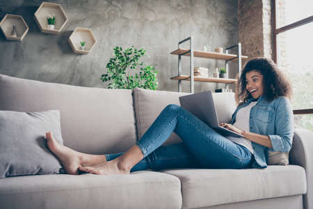 Profile photo of pretty amazed dark skin curly lady hold notebook texting with friends read cool good news lying comfy couch casual denim outfit living room indoorsの写真素材