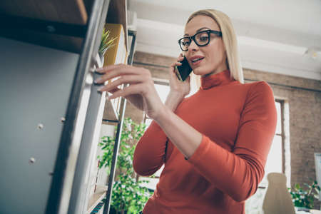 Low angle view photo of concentrated woman worker speak on smartphone do task take big folder from shelves wear red turtleneck in office loftの写真素材