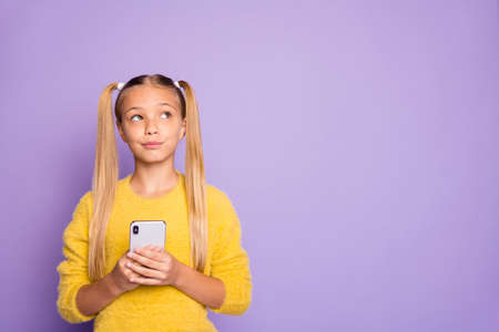 Photo of pensive contemplating looking girl staring into empty space to find idea with telephone in hands isolated in yellow sweater over pastel violet color backgroundの写真素材