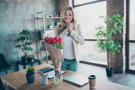 So cute. Photo of beautiful blond business lady came workplace looking affectionate fresh tulips bunch little postcard on table corporate unexpected 8 march surprise in officeの写真素材