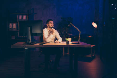 Photo of thoughtful dreamy serious man waiting for his shift to be over having done all necessary work bored sitting at desktopの写真素材