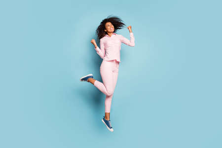 Full length photo of amazing dark skin lady jumping high celebrating cool victory triumphing wear pink shirt trousers sneakers isolated blue color backgroundの写真素材