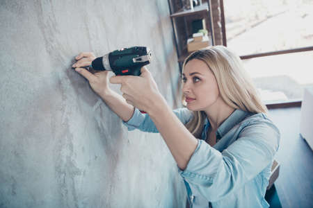 Profile side view portrait of her she nice attractive pretty lovely focused wavy-haired girl making hole in grey wall with drill at modern industrial loft brick style interior room indoorsの写真素材