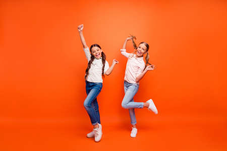 Full body photo of funky crazy two girlfriends have fun on fall weekends raise hands fists hold arms pigtails wear white shirt sneakers isolated orange color backgroundの写真素材
