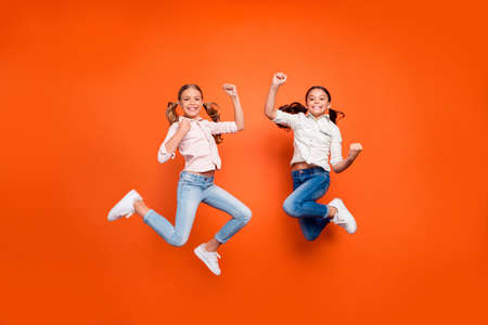 Full length photo of positive cute two children girls jump celebrate victory in school contest raise fists show strength wear white shirt denim jeans isolated orange color backgroundの写真素材