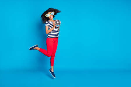 Full length turned body size photo of cheerful excited jumping girlfriend in striped t-shirt jumping with star shaped glasses isolated near empty space with disco ball vivid color blue backgroundの写真素材