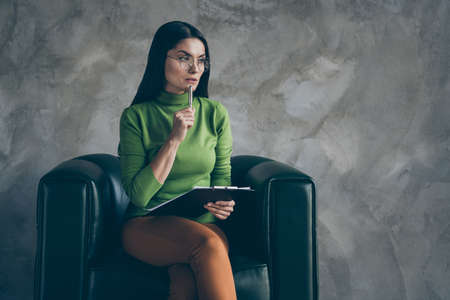 Photo of serious confident woman in orange pants touching chin with pen holding clipboard looking...